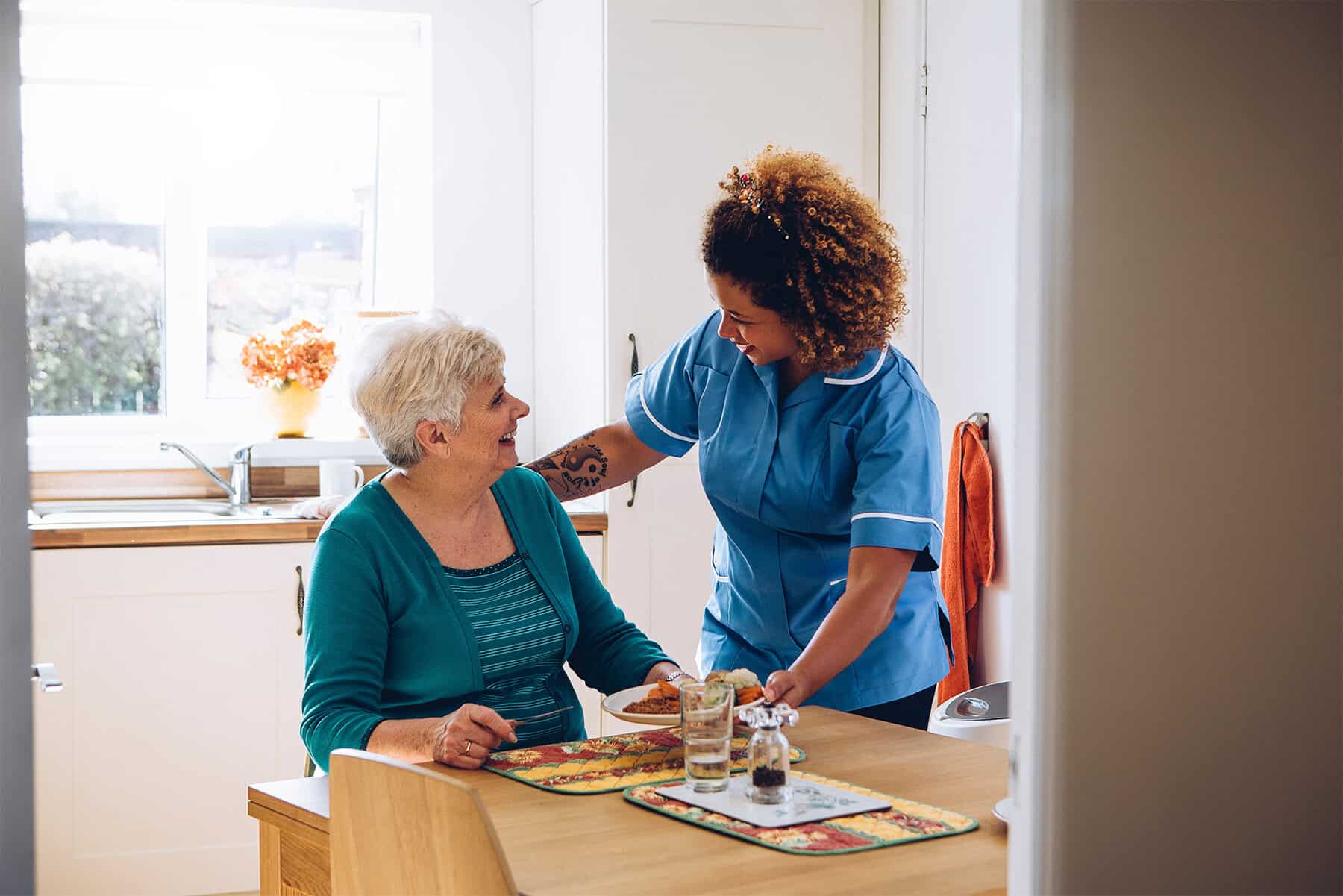 An elderly woman receiving compassionate home health care from a professional caregiver in a bright, welcoming kitchen setting.