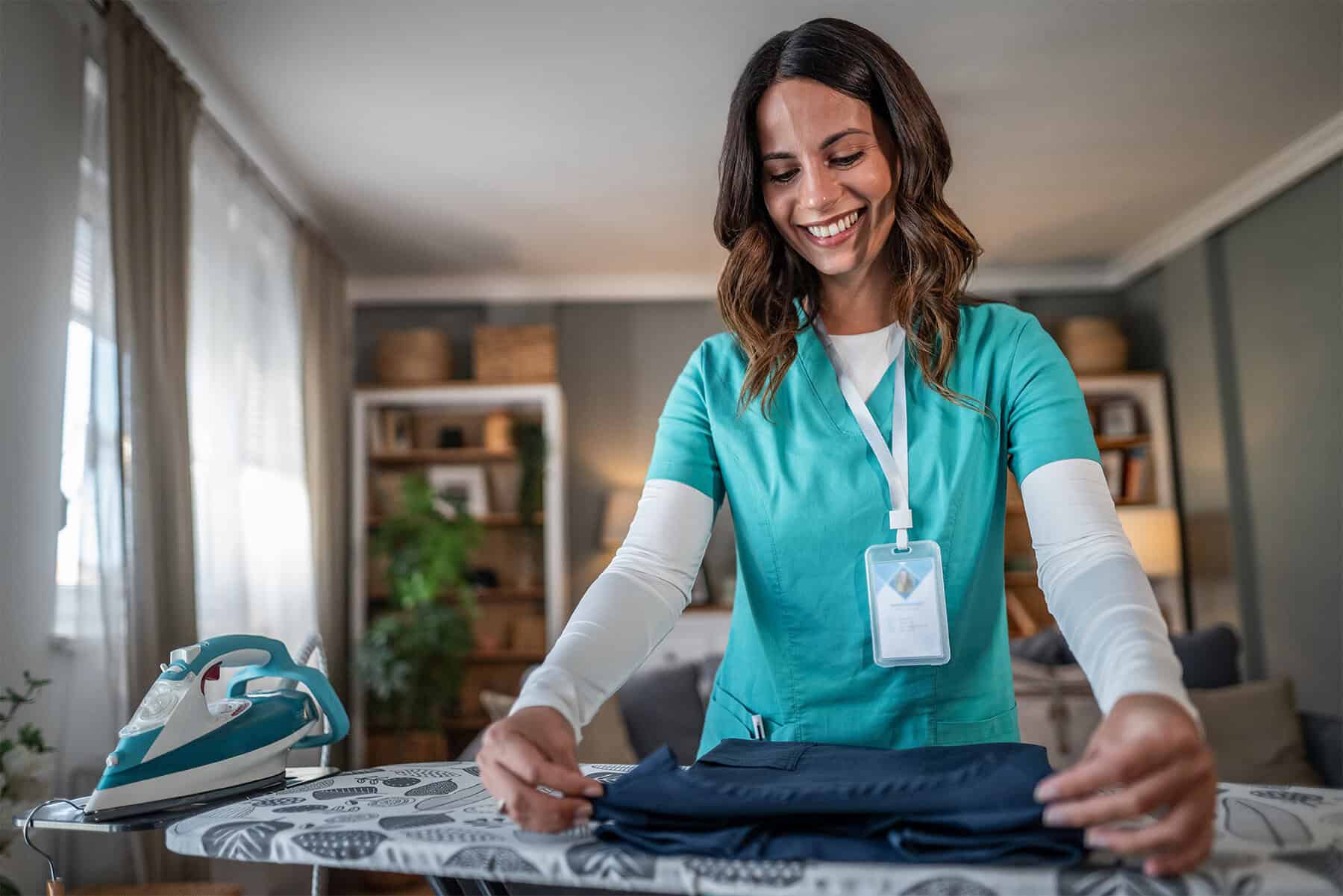 Friendly home health caregiver in scrubs folding laundry with an iron and ironing board, providing in-home care services for seniors and patients, emphasizing compassionate healthcare support.